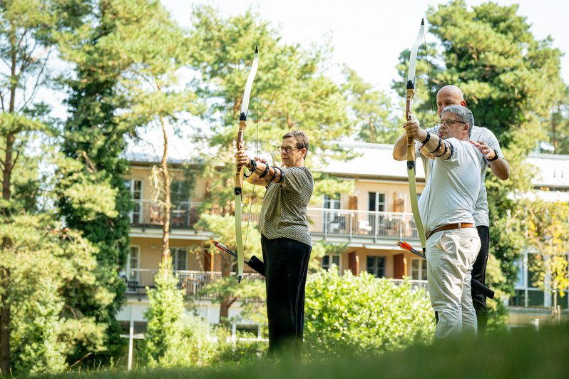 2 Personen üben Bogenschießen im Freien, weitere Person erklärt