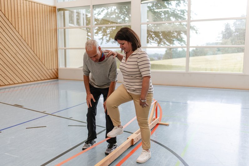 Frau balanciert mit Unterstützung eines Therapeuten auf einem Holzbalken in einer Turnhalle