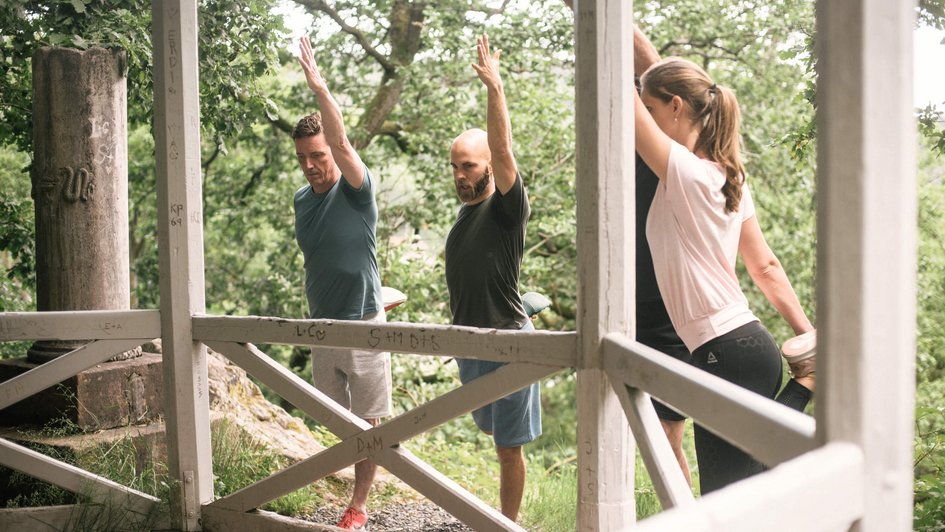 Vier Patienten stehen im Wald um Holzpavillon und führen Dehnübungen durch.