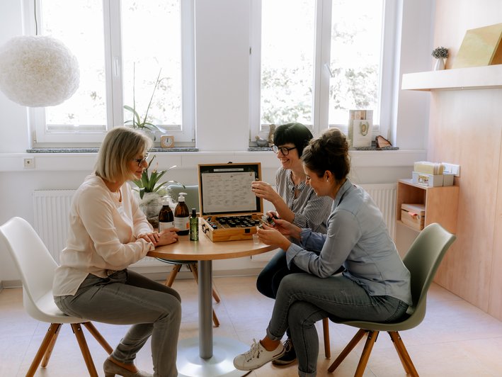 3 Frauen sitzen um einen kleinen Tisch herum, auf denen in einem Kasten kleine Fläschen mit Aromen sind