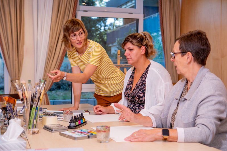 2 Frauen sitzen am Tisch, weiße Blätter vor Ihnen, eine Menge Malutensilien stehen herum, dritte Frau erklärt was zu tun ist