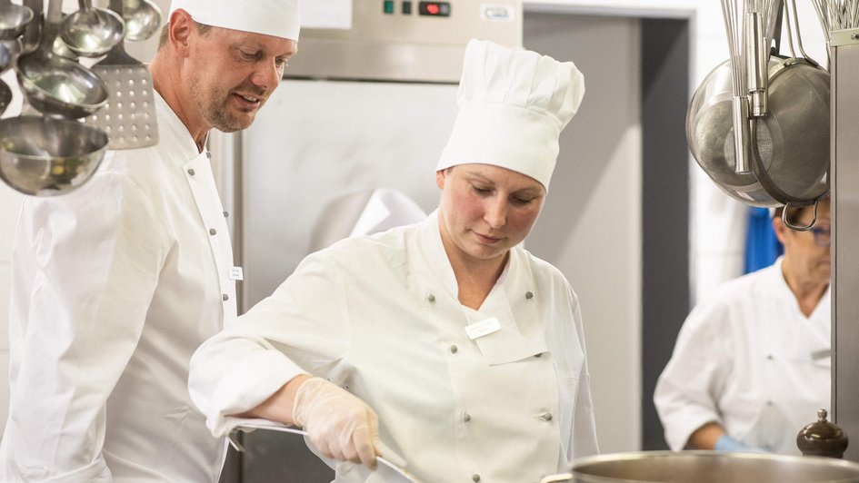 Ernährung Koch und Köchen stehen in der Klinikküche. Der Koch schaut der Köchin bei Ihrem Tun über die Schulter.