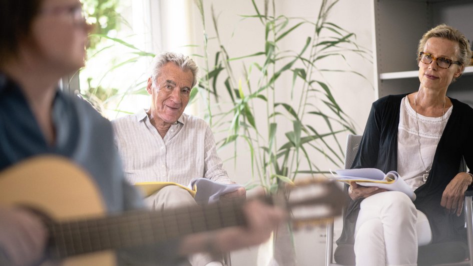 Therapieangebot Fachklinik Berlin Brandenburg  Zwei Frauen und ein Mann sitzen in einem Raum. Eine der Frauen spielt Gitarre.