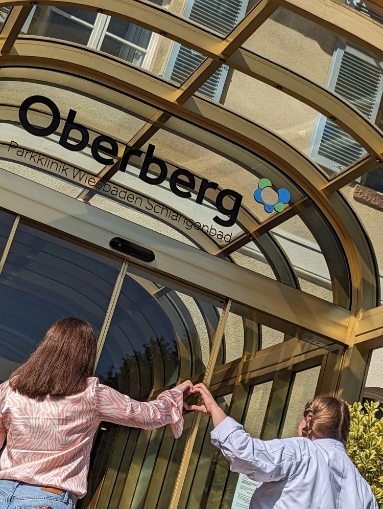2 junge Frauen sthen vor dem Eingang der Oberberg Parkklinik Wiesbaden Schlangenbad, bilden ein Herz mit ihren Händen