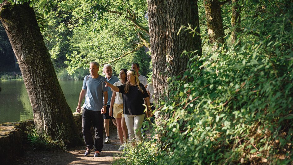 Eine Gruppe von Menschen geht durch den Wald spazieren, im Hintergrund ein See.  