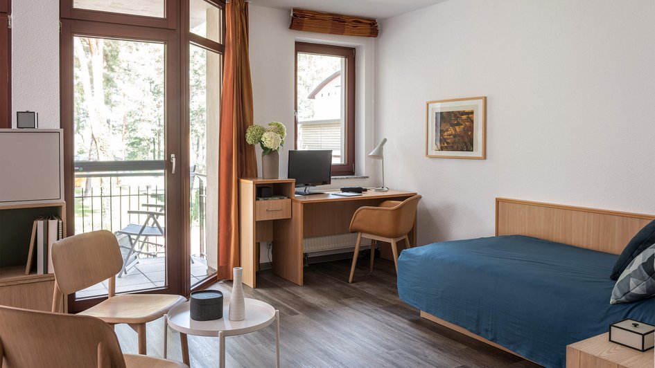 Räumlichkeiten der Klinik Berlin Brandenburg  Möbliertes Patientenzimmer mit Bett, Schreibtisch, Sitzgelegenheiten und Balkon.
