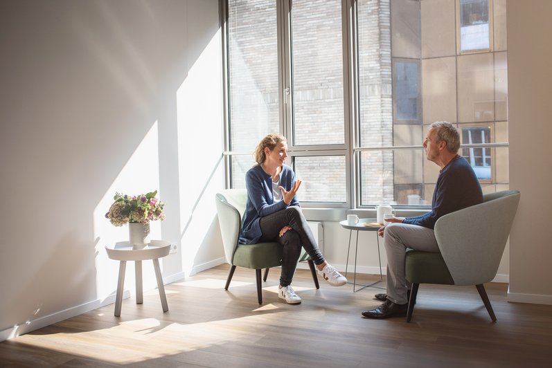 Personen sitzen in Sesseln und unterhalten sich, im Hintergrund eine große Fensterfront die viel Licht herein lässt