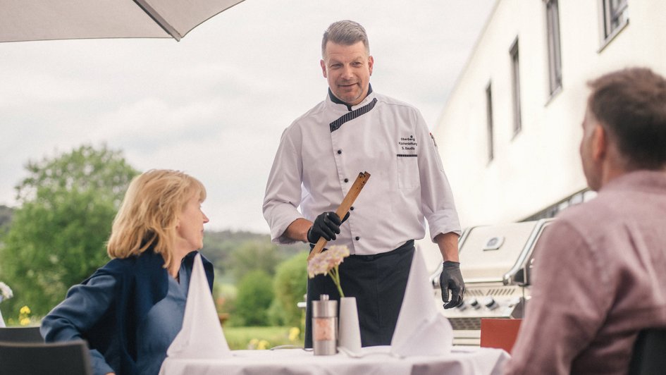 Koch mit Zange in der Hand an einem Tisch mit zwei Personen