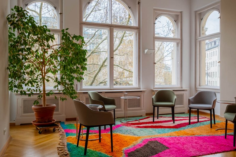 mehrere Stühle stehen im Kreis, großer bunter Teppich auf dem Boden, viel Licht durch große Fenster