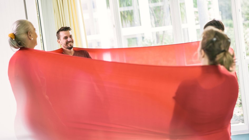 Therapieverfahren Vier Personen stehen sich gegenüber im Kreis, umwickelt von einem roten Tuch