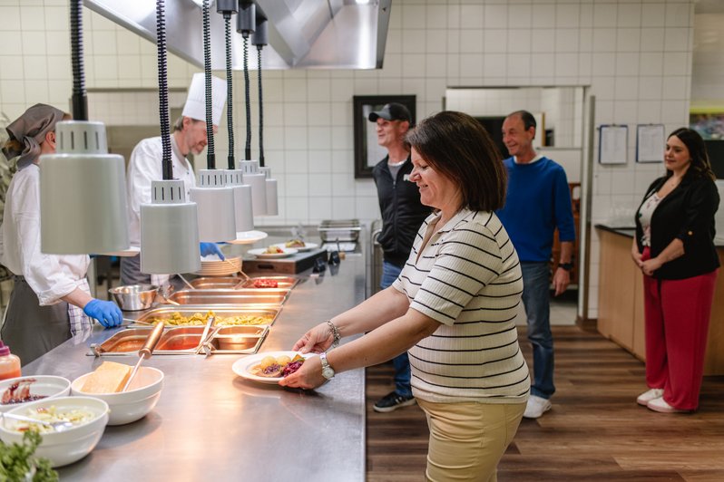 Menschen in der Essensausgabe, eine Frau nimmt ein frisch zubereitetes Gericht von der Theke entgegen