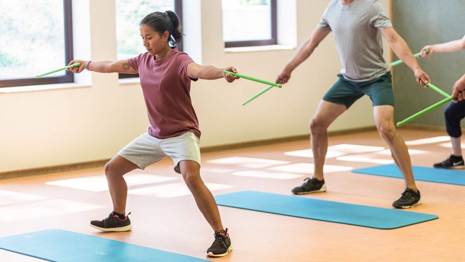 Therapieangebot Fachklinik Berlin Brandenburg Gruppe von Menschen steht in leicht gehockter Stellung hinter einer Gymnastikmatte. Sie halten Holzstäbe in ihren ausgestreckten Händen.