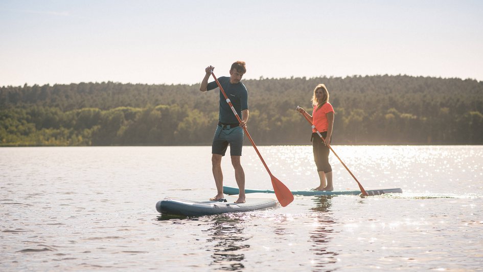 Freizeit und Umgebung Eine Frau und ein Mann laufen auf einem Steg und tragen Stand-up-Paddling-Boards. Im Hintergrund Schilf